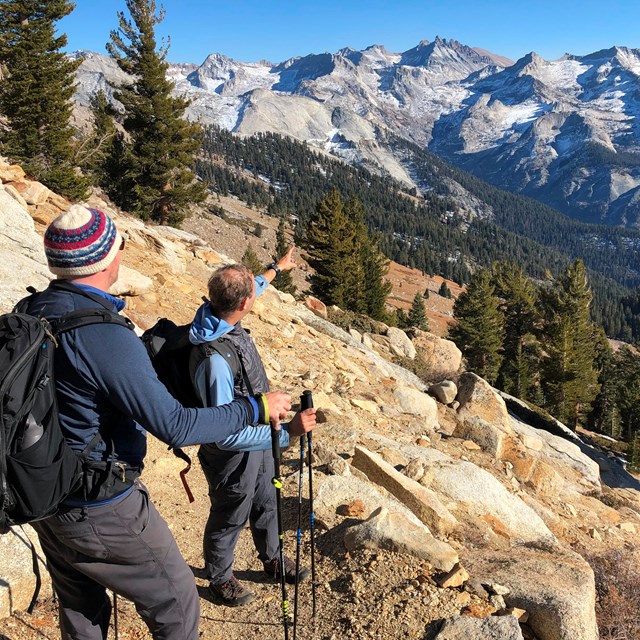 Two men in backpacks on a rocky trail look out over a vista of rugged mountains and conifers.