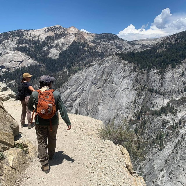 Hikers walk on a gravelly trail next to a steep cliff with rugged mountains in the background. 