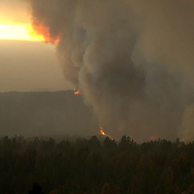 A view of gray smoke rising from a dark forest and a narrow horizontal band of daylight below clouds