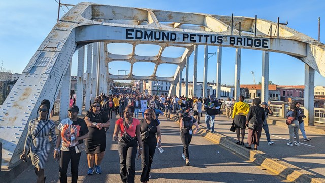 A crowd walking across a bridge with steel beams overhead that read "Edmund Pettus Bridge"