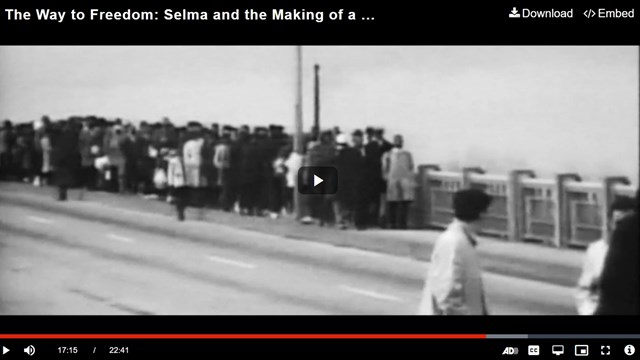 Still of a video showing black and white footage of a line of people walking across a bridge.