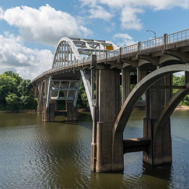 View from the side and below of a bridge spanning a river. A blue sky frames the background.