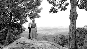 A black and white historical image showing a couple standing on a rock at an overlook