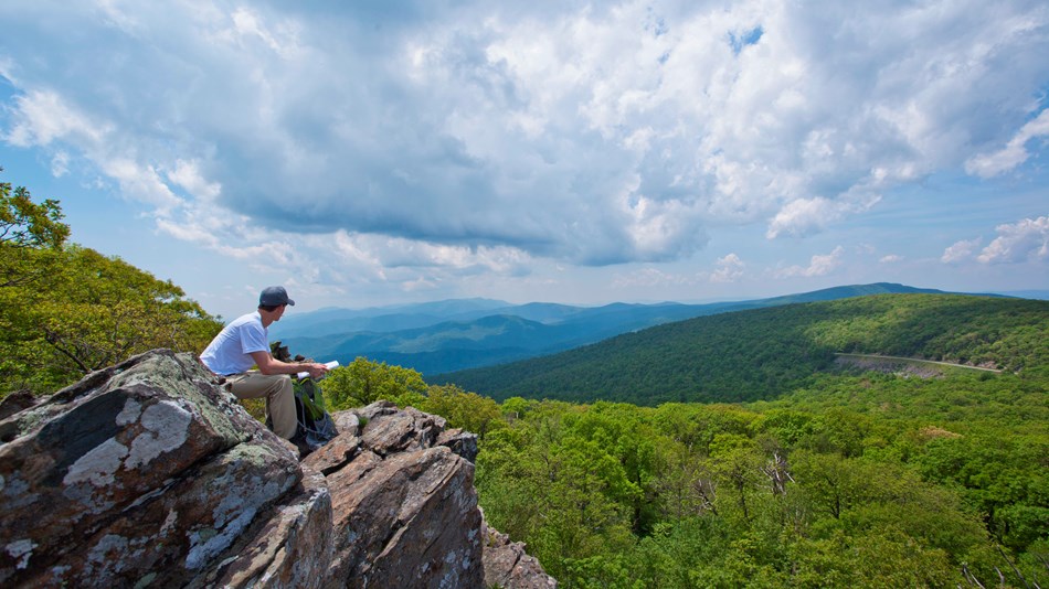 Hiker on large rock outcrop studies beautiful mountain ranges in the distance.
