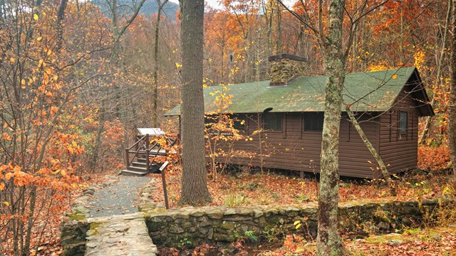 A stone bridge leads to a historic brown-sided building with a green roof and a chimney.