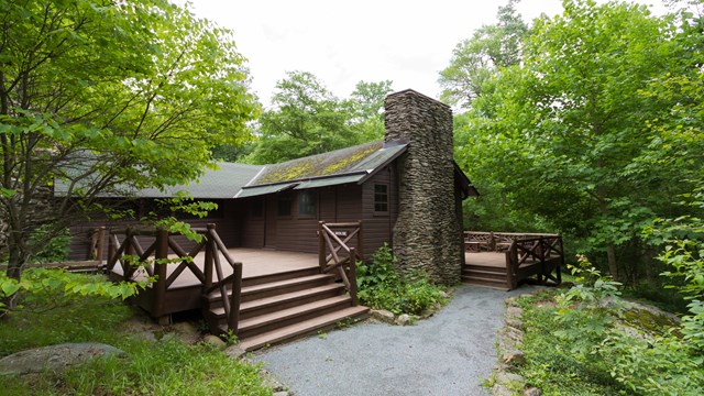 An historic cabin with brown siding, a chimney, and a large porch amidst a forest of green trees.