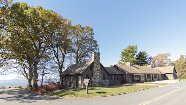 A road leads past a single-level stone building in front of blue mountains in a valley below it.