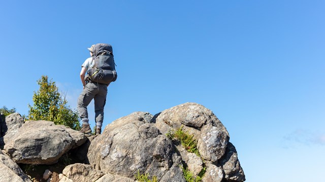 A male hiker wearing a backpack standing on a series of boulders against a brilliant blue sky.