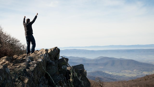A man stands on a rocky outcrop with his arms outstretched. A view of foothills is in the background