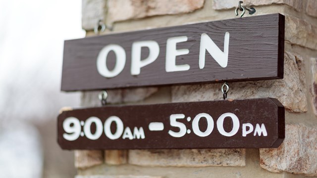 A brown open sign hanging from a stone wall. A small sign under it reads: 9 a.m. to 5 p.m.