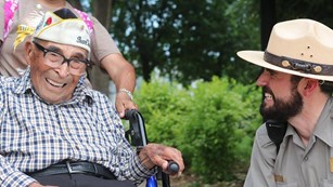 An elderly man in a wheelchair with a military hat smiles with a male park ranger
