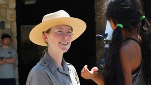 A female park ranger smiles at a young girl looking at a telescope.