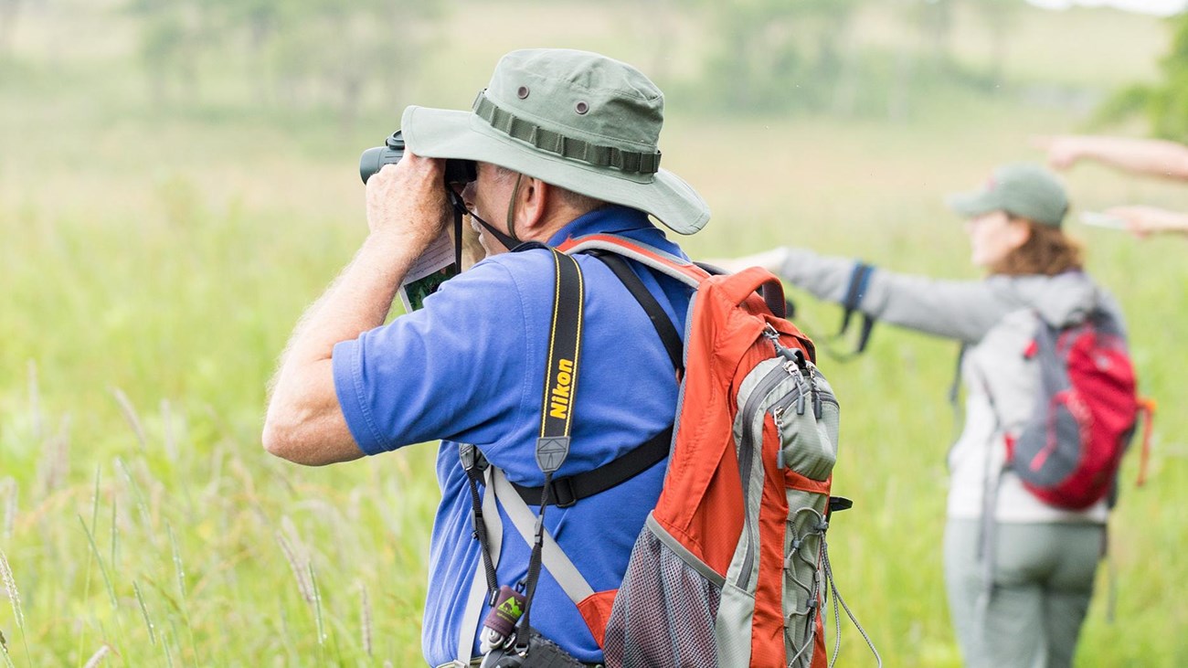 A man with binoculars looking to the distance with people in the background are pointing.