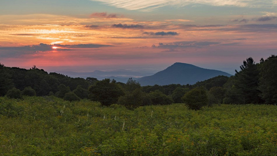 A sun rises behind a green mountain overlook.