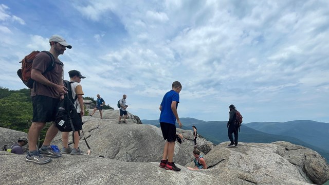 A large group of people hiking on a mountain top full of boulders and rocks.