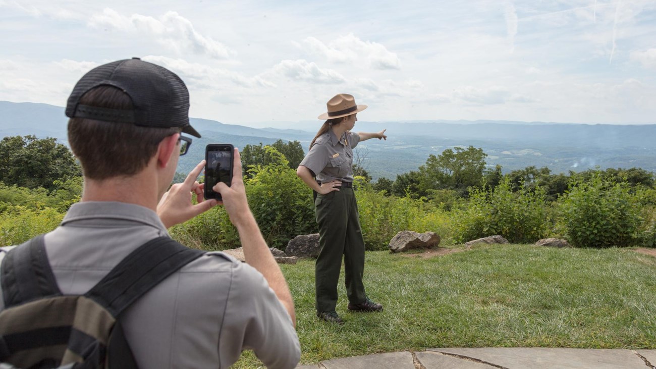 A Park Ranger takes a video of a fellow Park Ranger with their phone at a view of mountains.
