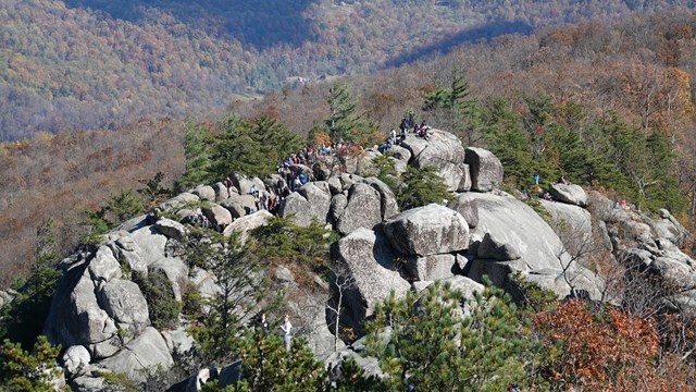 A large group of people hiking on a mountain top full of boulders and rocks.