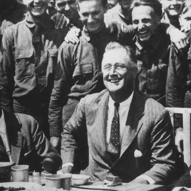 A black and white image of President Hoover sitting at a dinner table with a group of CCC boys.