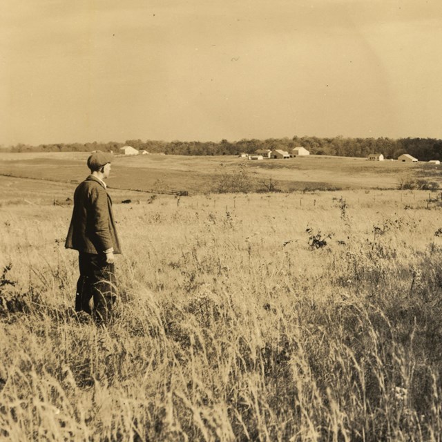 A sepia-toned photograph of a man standing in a field, looking off in the distance.