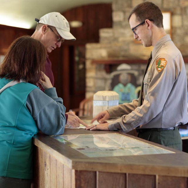 A Park Ranger stands behind a desk at a visitor center and answers questions for visitors.