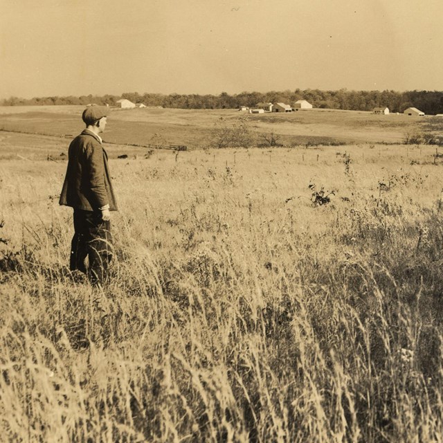A sepia tone historical image of a man looking across a field to mountains in the distance.