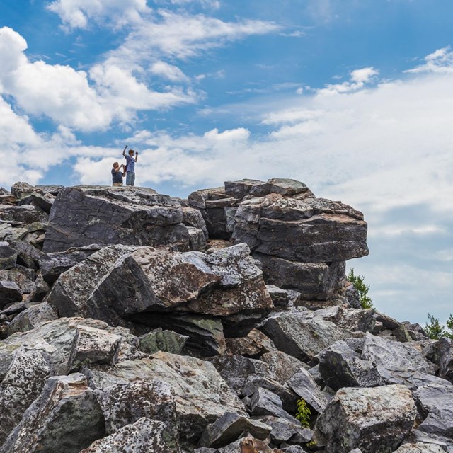 Two hikers stand on top of a large pile of rocks, looking out over a mountainous view.