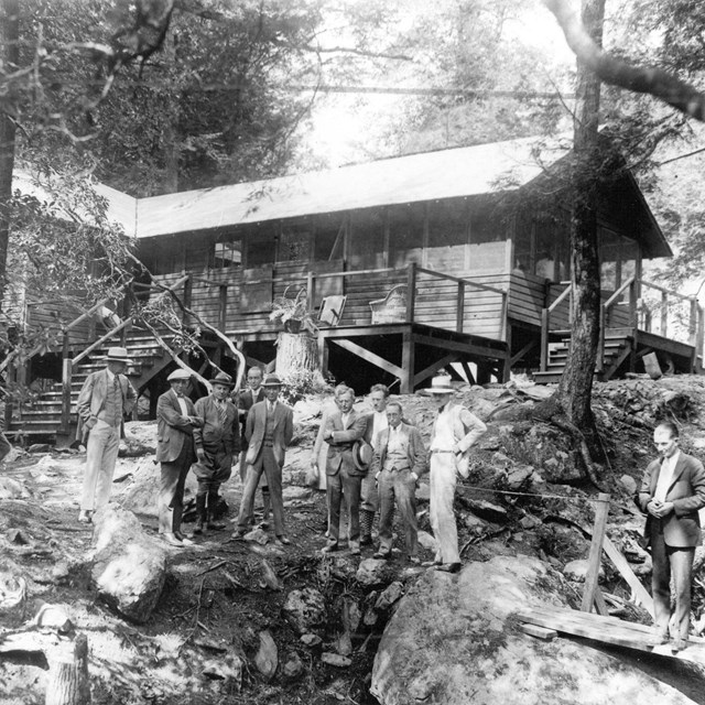 A black and white image of a group of reporters standing outside of a rustic building, Camp Hoover.