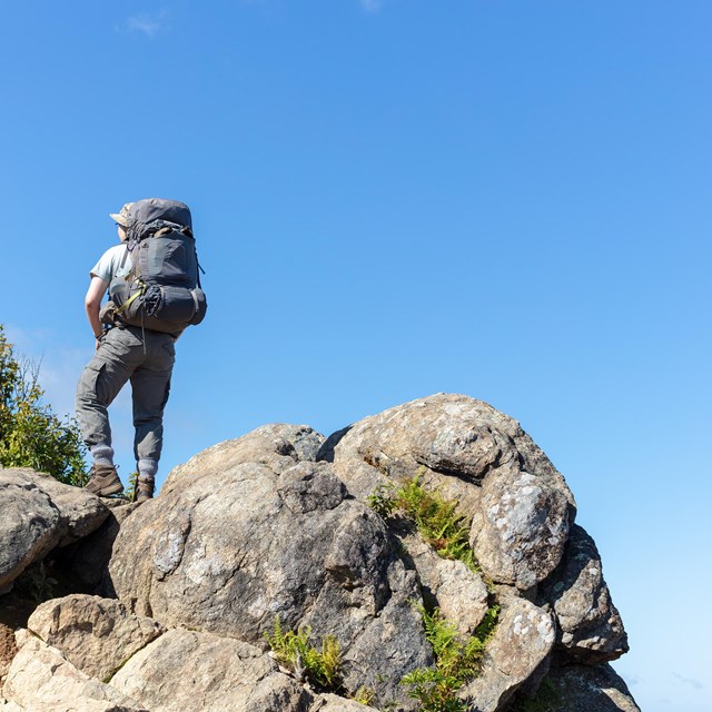 A male hiker wearing a backpack standing on a series of boulders against a brilliant blue sky.