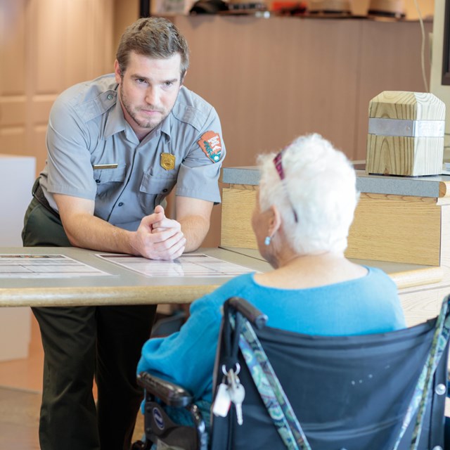 A park ranger leans over a desk to answer a question from a woman in a wheel chair.