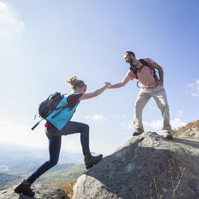 A man extends his hand to help a woman climb up a large rock.