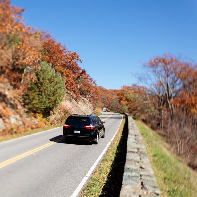 A black car driving down the a road next to a rock wall lined with fall colors of red and orange.
