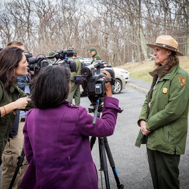 A female park employee stands calmly in front of a group of reporters with video cameras.