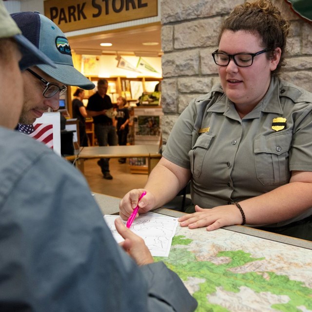 A female park ranger shows two visitors a map on a visitor center desk.