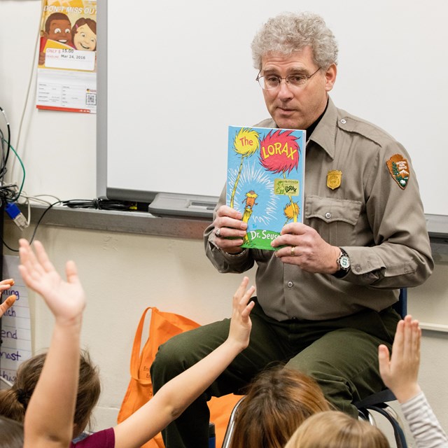 A park ranger holding up a book in a classroom full of children with their hands raised