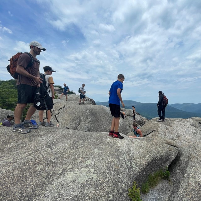 A large group of people hiking on a mountain top full of boulders and rocks.
