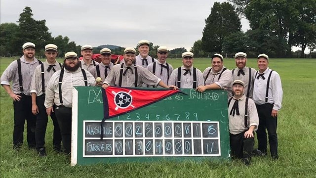 Base ball players in period uniforms stand behind a score board.  