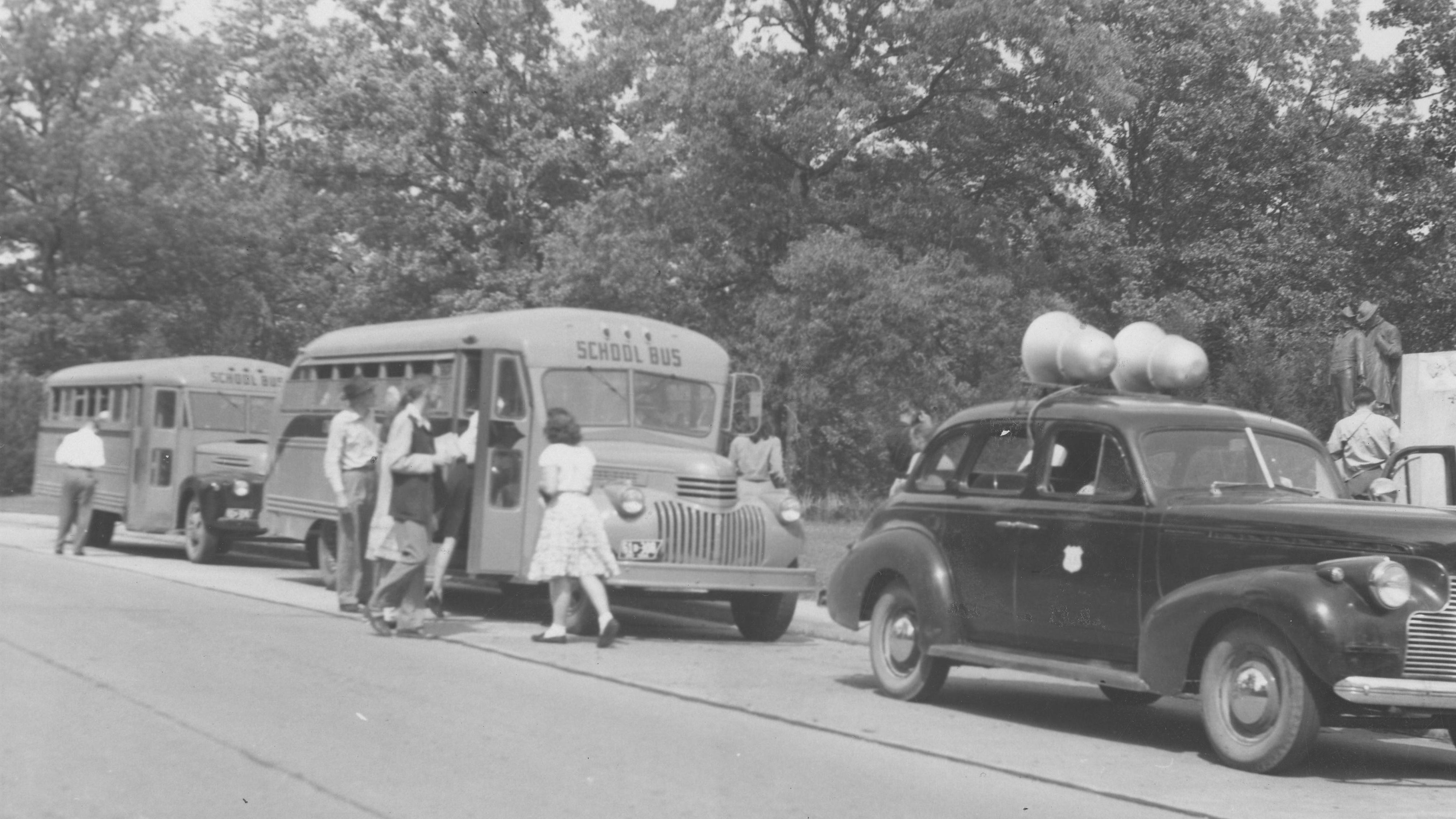 Black‑and‑white photograph shows two school buses parked on a roadside next to a wooded area