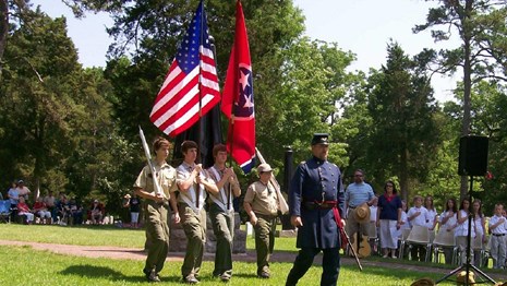 Young men in military uniforms march across a green grassy field before a crowd of people.