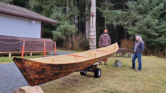 A dugout canoe on a trailer on a lawn. Two men and a totem pole near a forest beyond the canoe.