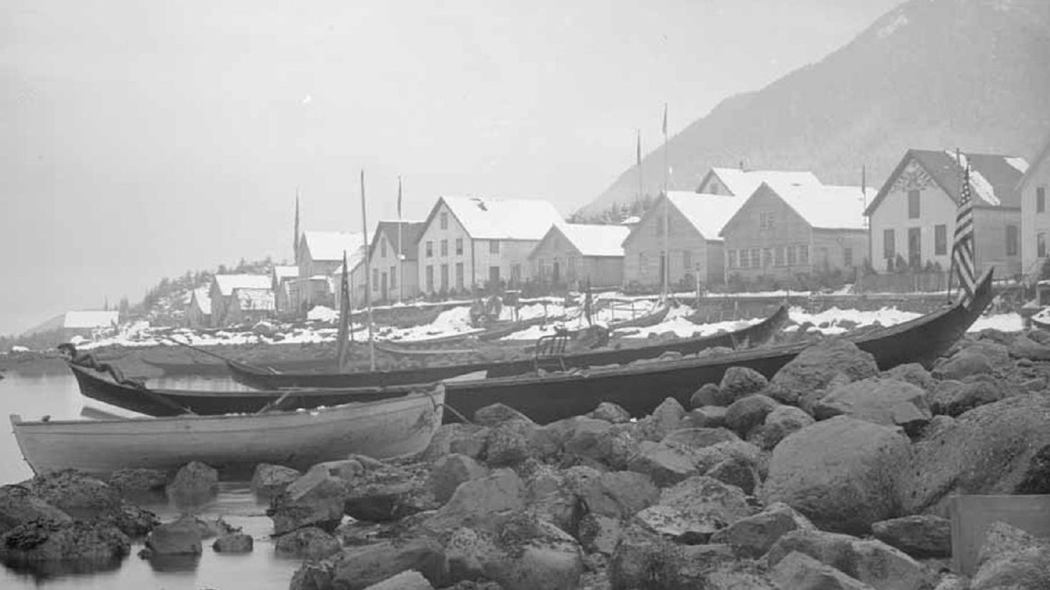 A historical photo of traditional dugout canoes on a rocky shore in front of houses lining the coast