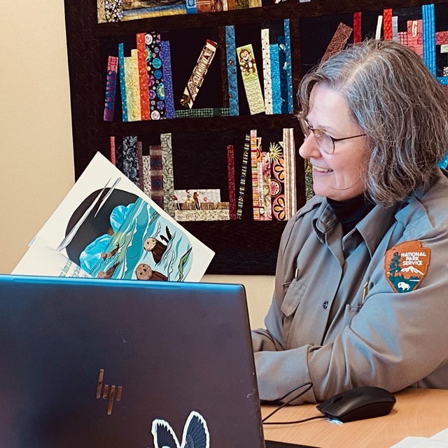 A female park ranger holds a storybook to a laptop computer.