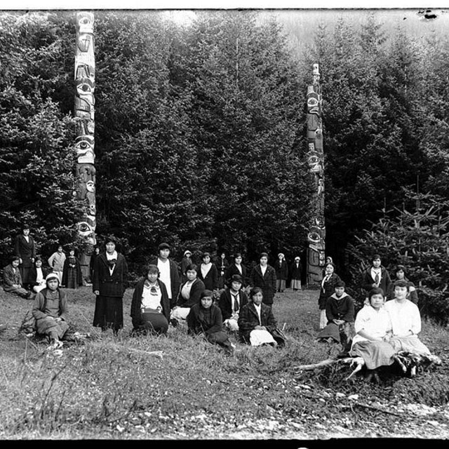 black and white photo of a group of people sitting in the grass around two totem poles in the forest