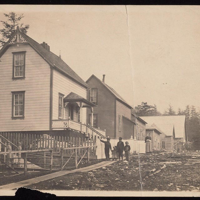 old photograph of a row of wooden houses with children and a women walking on a wood plank sidewalk 