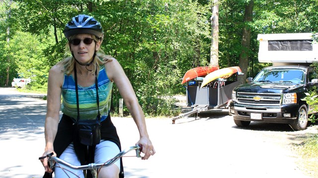 A woman rides her bike past a campsite with kayaks and a pickup camper.