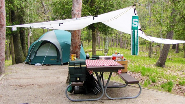 A tent, picnic table with red and white checked cloth, and sun shield make an inviting campsite.