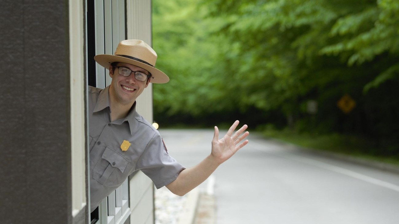 A male ranger in a flat hat leans out the window of a fee kiosk and waves.