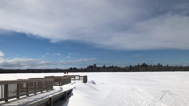 View of a snow-covered frozen lake and wooden dock under a blue sky with ice fishing tents