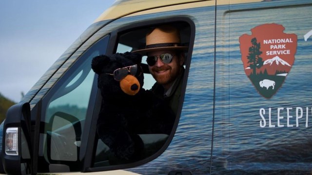 A park ranger smiles in a colorful van while holding a stuffed animal bear