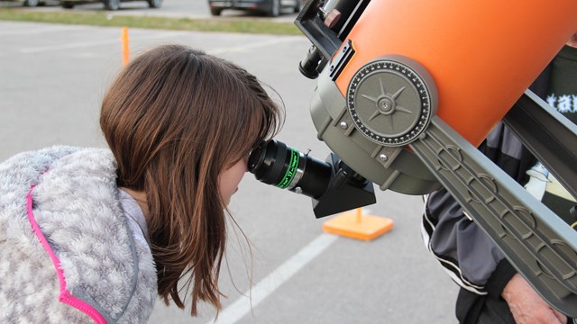 A girl looks through the viewfinder of a large, orange telescope.