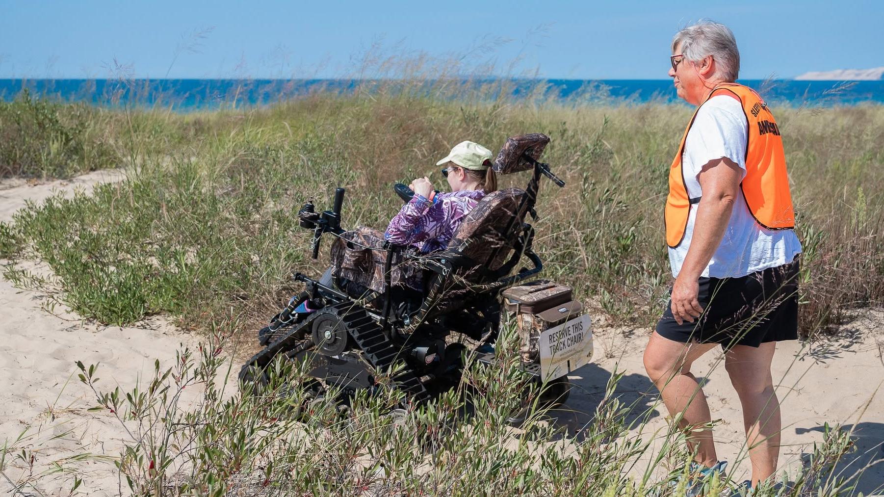 A person is a track wheelchair drives on the sand with a person walking behind